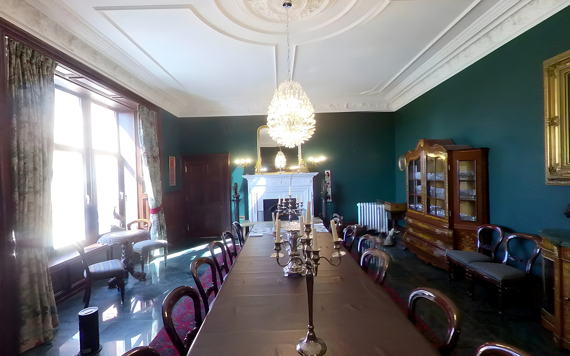 The same dining room in April 2025: green walls, white plaster ceiling, chandelier and long table, with sun through the garden window