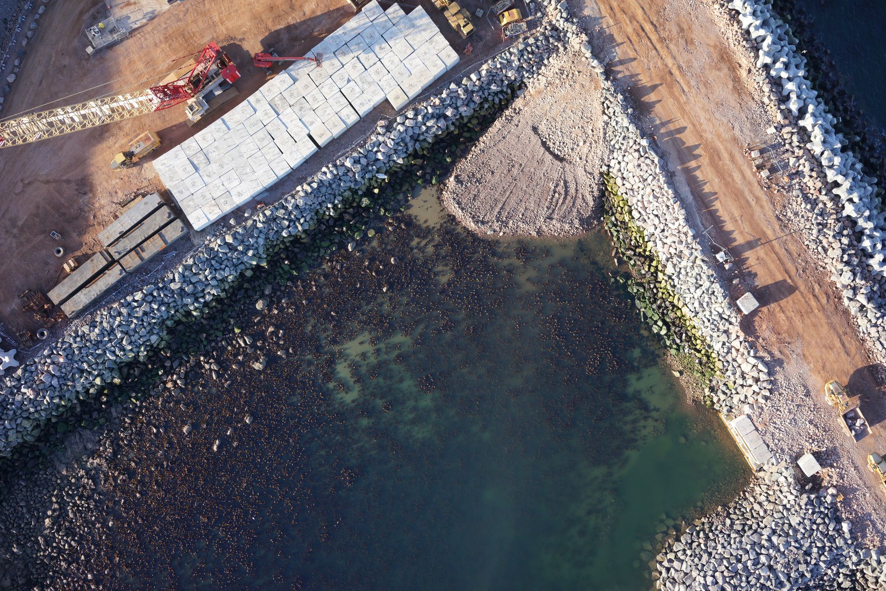 Straight-down drone photo of a harbour under construction, with the precast block yard, crane lifting zone, rock armour, quay infill, concrete armour units and the haul road marked